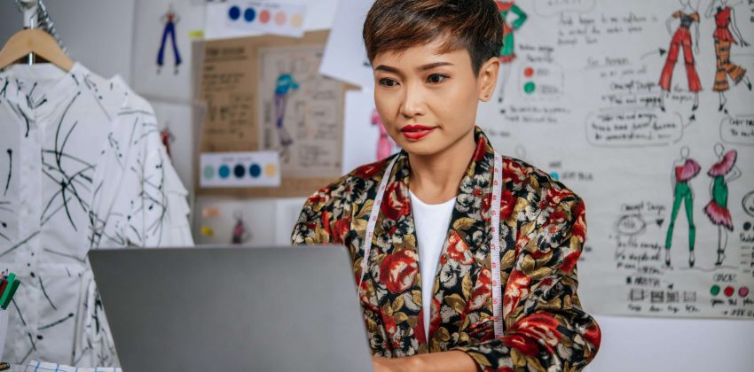 Young beautiful designer use laptop computer to searching idea for design clothes, Tape measure on her neck, clothes sketch on board in background